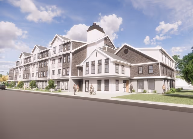 Exterior view of a large multi-story residential building with pitched roofs, a sidewalk with a few people, and a partly cloudy sky.