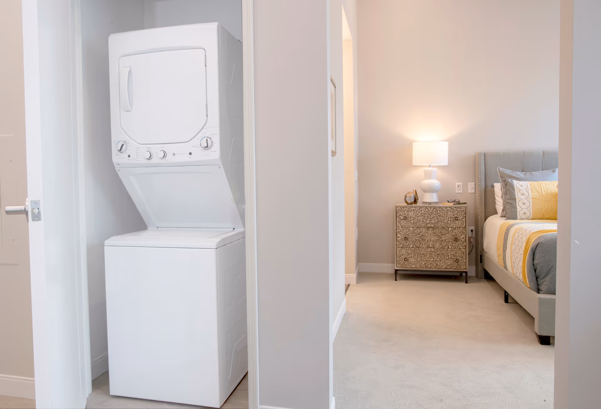 A white stacked washer and dryer unit is placed in a small laundry closet with an open door. Adjacent to the laundry area is a bedroom with a beige carpet, a bed with a gray upholstered headboard, and a nightstand with an ornate design. A white table lamp is on the nightstand, casting a warm light on the wall.
