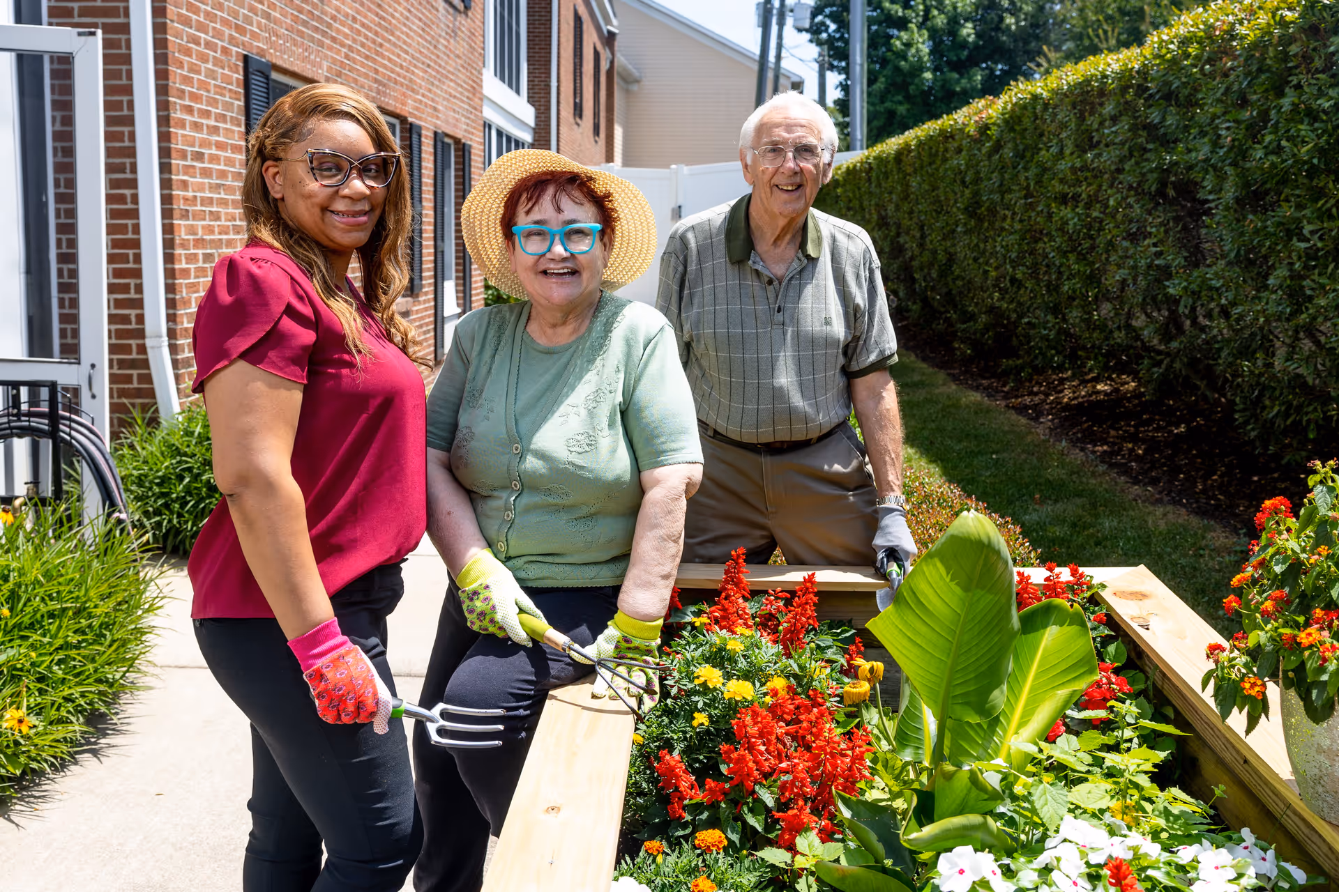Three people, two elderly individuals and a younger woman, are gardening together outdoors next to a raised flower bed filled with colorful flowers and green plants. They are smiling and wearing gardening gloves, with a brick building and a hedge visible in the background.