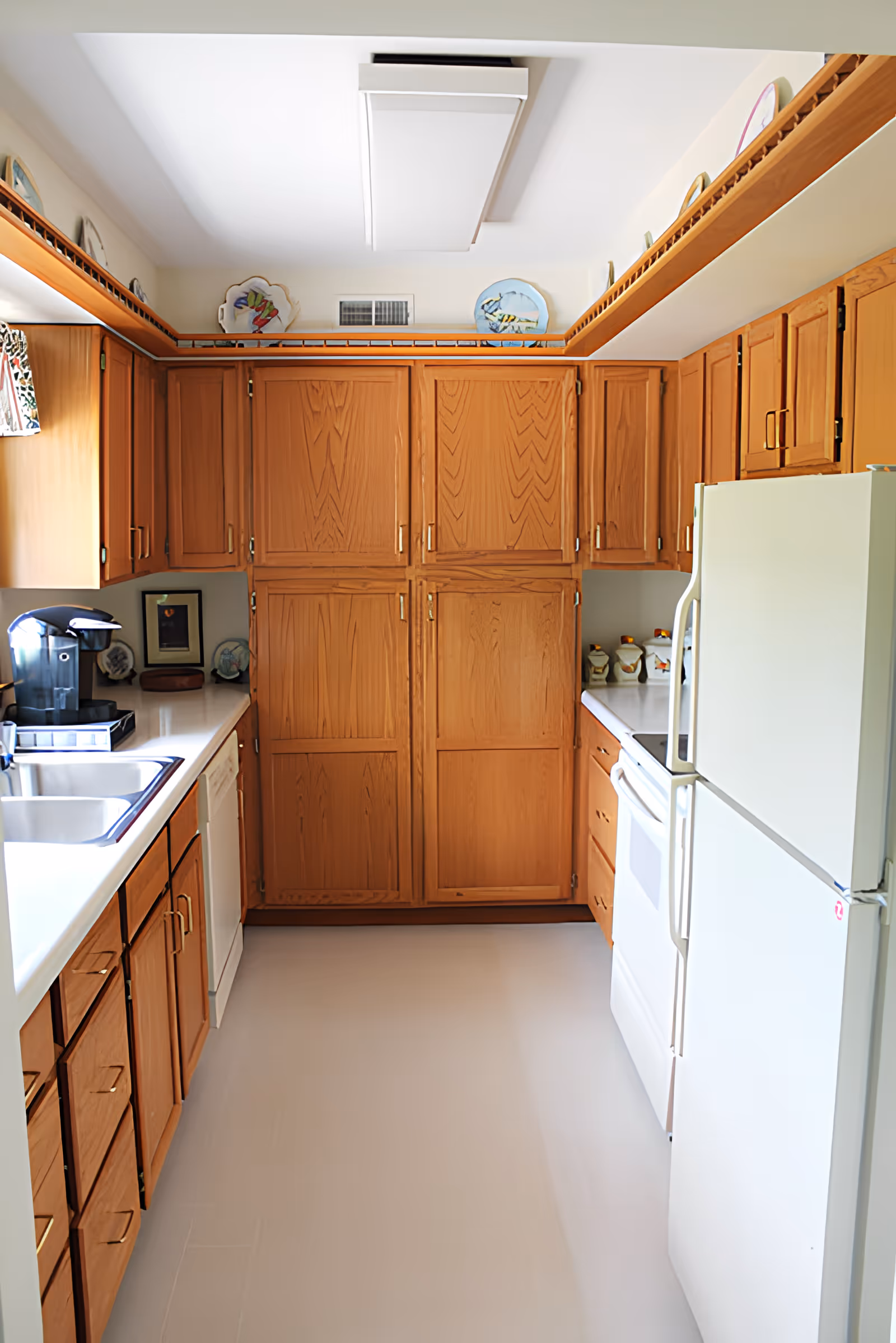 A bright kitchen with wooden cabinets, white countertops, a white refrigerator, a white stove, a dishwasher, a double sink, and a coffee maker. Decorative plates are displayed on a shelf near the ceiling.