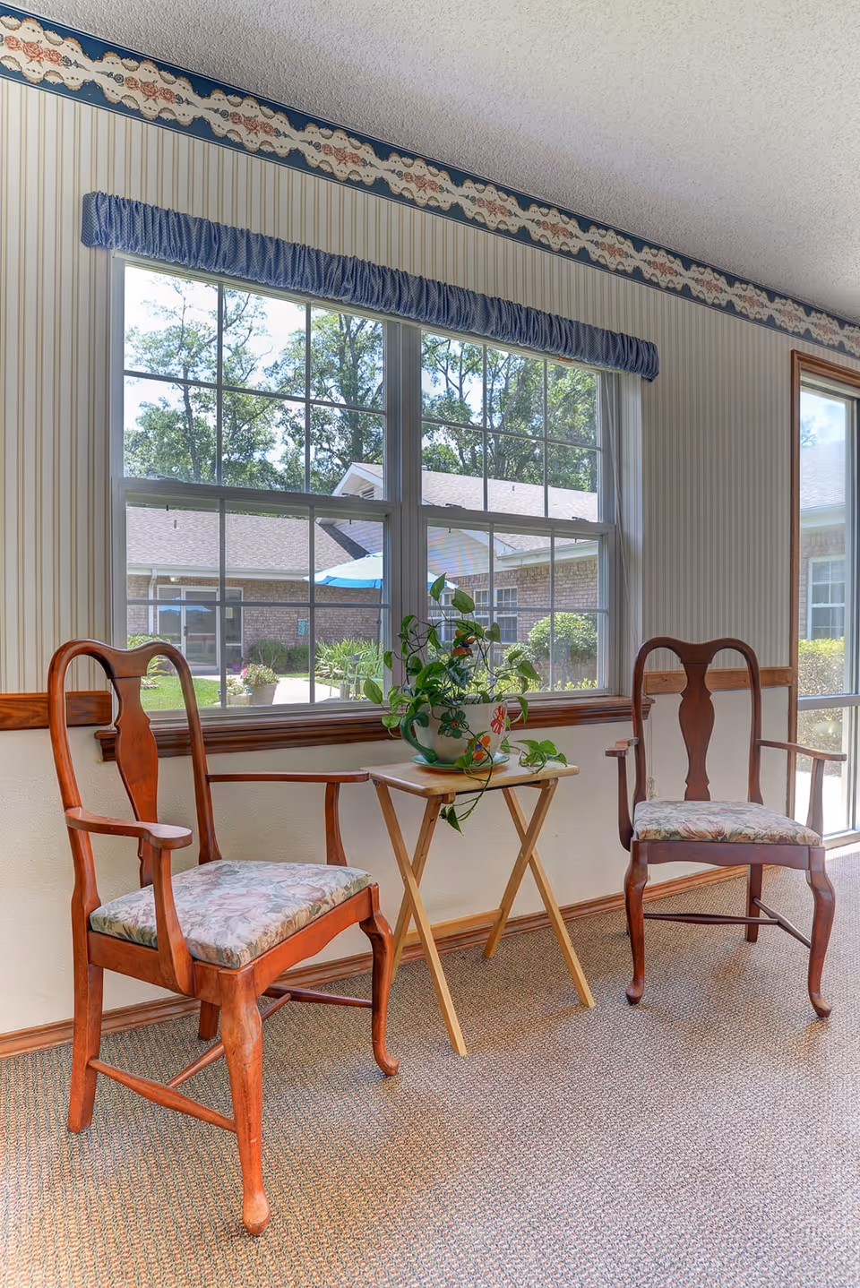 Two wooden armchairs with floral cushioned seats are positioned on either side of a small wooden folding table holding a potted plant with green leaves. The setting is indoors with a large window behind the chairs showing an outdoor courtyard with trees, a blue umbrella, and a brick building. The room has striped wallpaper with a floral border near the ceiling and carpeted flooring.