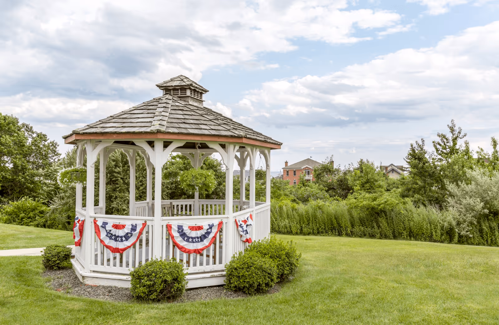 A white wooden gazebo decorated with red, white, and blue patriotic bunting, situated on a well-maintained grassy lawn with bushes around its base. Trees and houses are visible in the background under a partly cloudy sky.