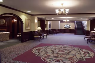 Spacious interior lobby area with patterned carpet, several chairs and tables along the walls, a chandelier hanging from the ceiling, and a staircase leading to an upper floor. The space is warmly lit and decorated with framed artwork and lamps.
