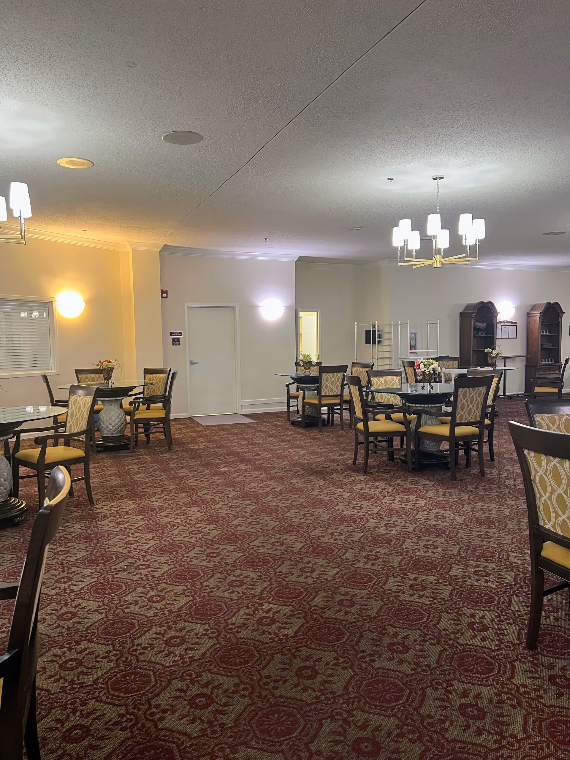 A spacious dining room with multiple round tables and chairs arranged neatly. The room has patterned red and beige carpet, cream-colored walls, and soft lighting from wall sconces and chandeliers. There are decorative flower arrangements on the tables and wooden cabinets along the back wall.