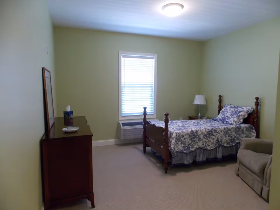 A simple bedroom with light green walls, a single wooden bed with blue and white floral bedding, a wooden dresser with a tissue box and a plate, a window with blinds, a small nightstand with a lamp, and a beige armchair.