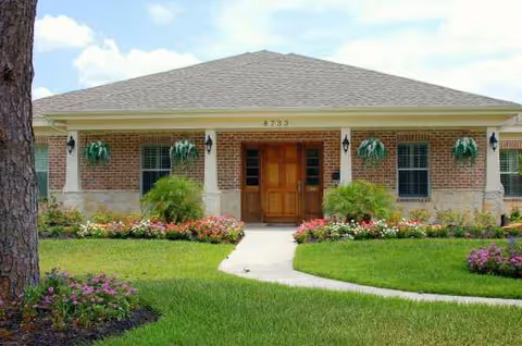 Single-story brick senior living front with a wooden door, manicured lawn, and hanging plants.