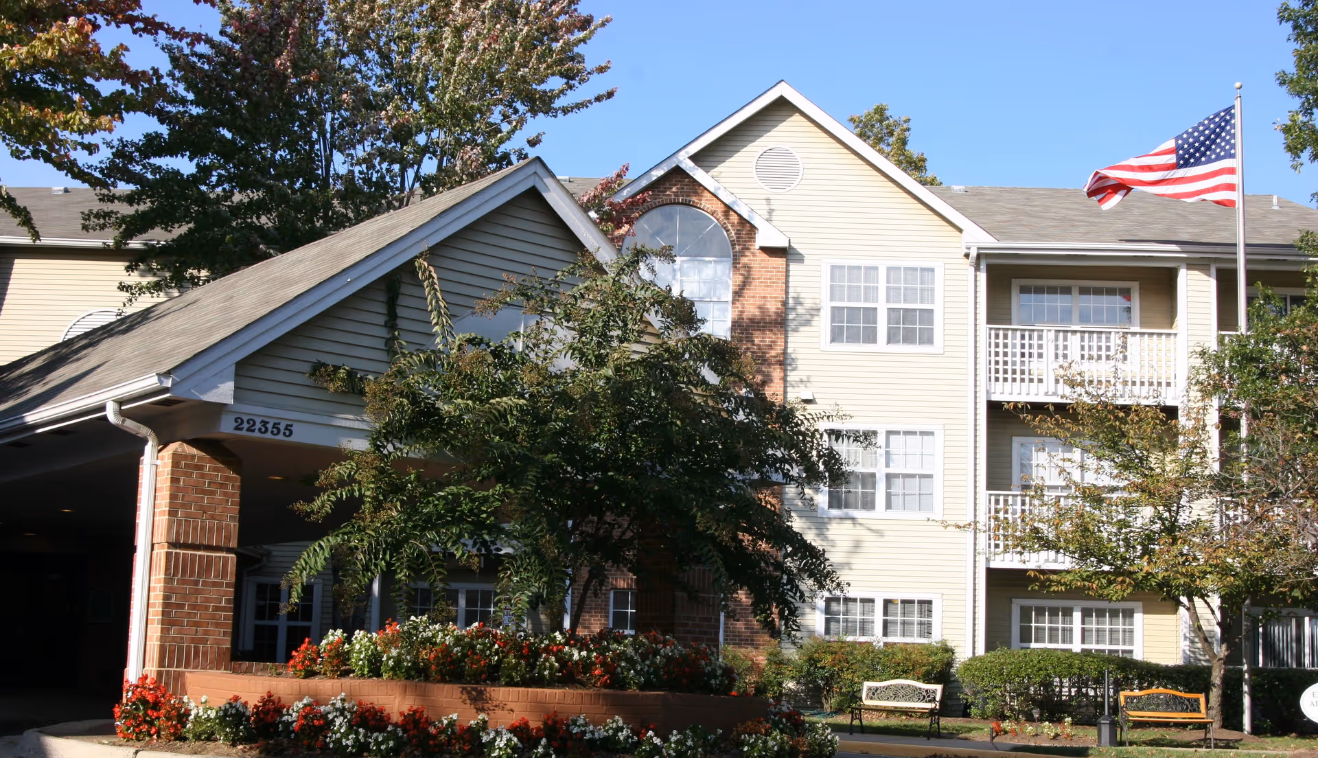 Exterior view of a multi-story retirement community building with beige siding and brick accents. The entrance has a covered driveway with the number 22355 displayed. There are trees, flower beds, and benches in front of the building, and an American flag is flying on a flagpole to the right.