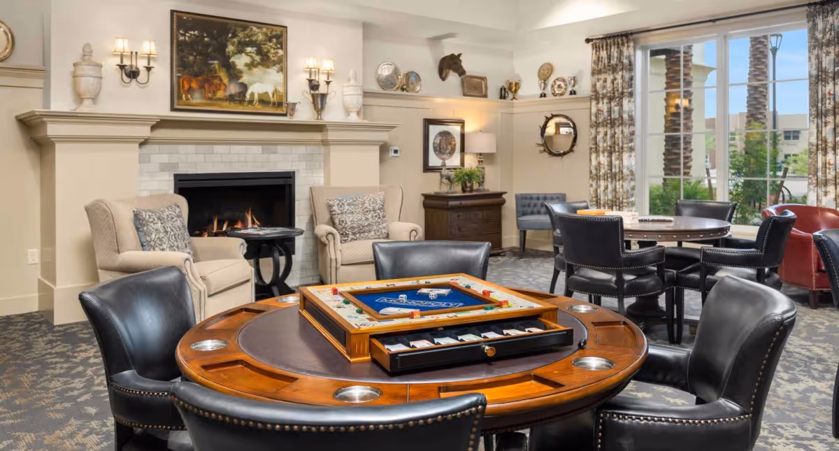A cozy common area in a senior living community featuring a round wooden game table with chairs around it, a fireplace with a painting above it, two beige armchairs with patterned pillows, and large windows with floral curtains letting in natural light.