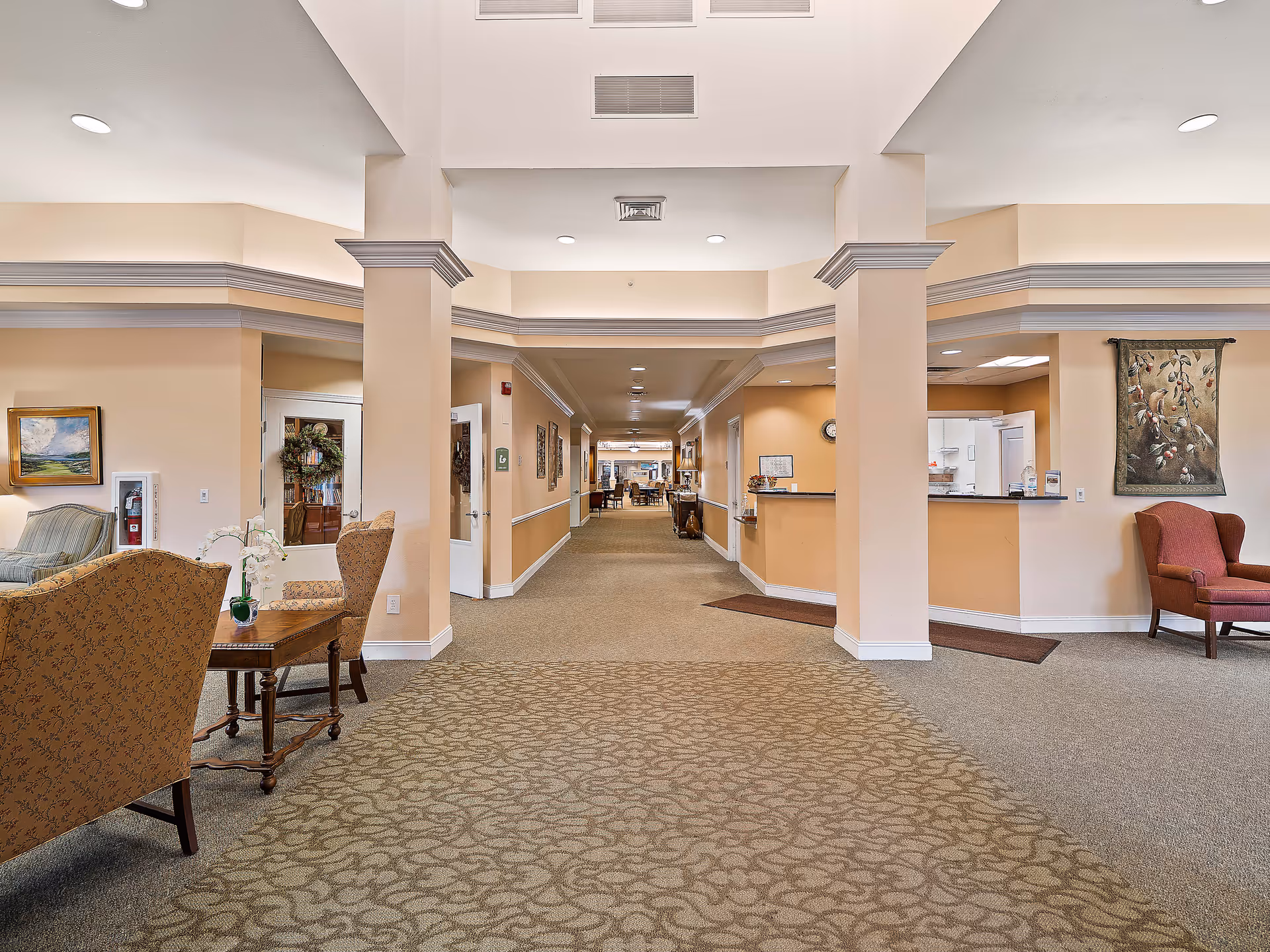 Interior view of a senior living facility hallway with beige walls and carpeted floors. There are seating areas with upholstered chairs and small tables on either side. The hallway leads to a common area with more seating and tables. The space is well-lit with recessed ceiling lights and decorated with framed artwork and a tapestry.