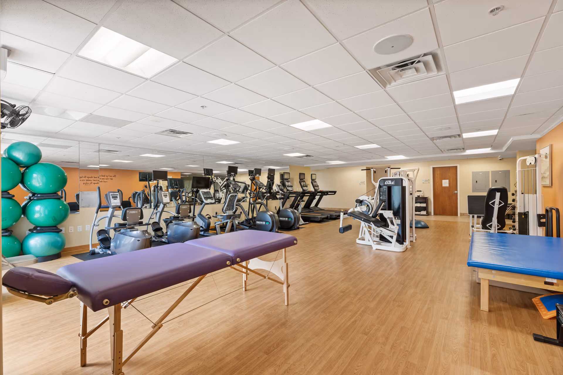 A spacious fitness room with wooden flooring and a white ceiling with recessed lighting. The room contains various exercise equipment including treadmills, stationary bikes, elliptical machines, and strength training machines. There are two padded tables, one purple and one blue, likely for physical therapy or stretching. On the left side, there is a rack holding several large green exercise balls. A large mirror covers the wall behind the exercise machines, reflecting the room.