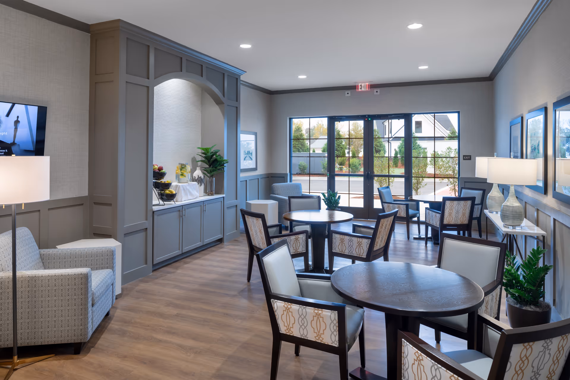 A bright and modern common area with round tables and cushioned chairs arranged for socializing or dining. The room features wood flooring, neutral-colored walls with wainscoting, large windows and glass doors letting in natural light, and decorative plants. There is a built-in cabinet with fruit and water dispensers, and lamps providing additional lighting.