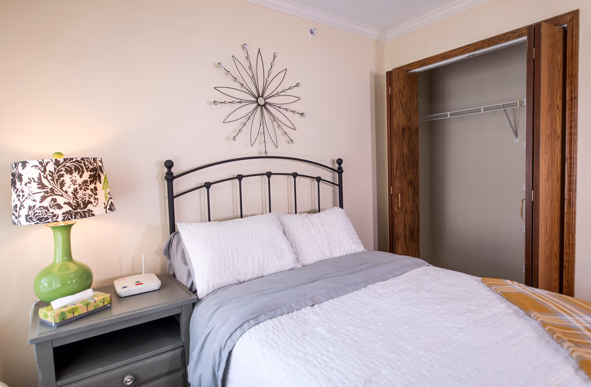 A tidy bedroom with a made bed, metal headboard, decorative wall art, a nightstand with a green lamp and tissues, and an open closet.