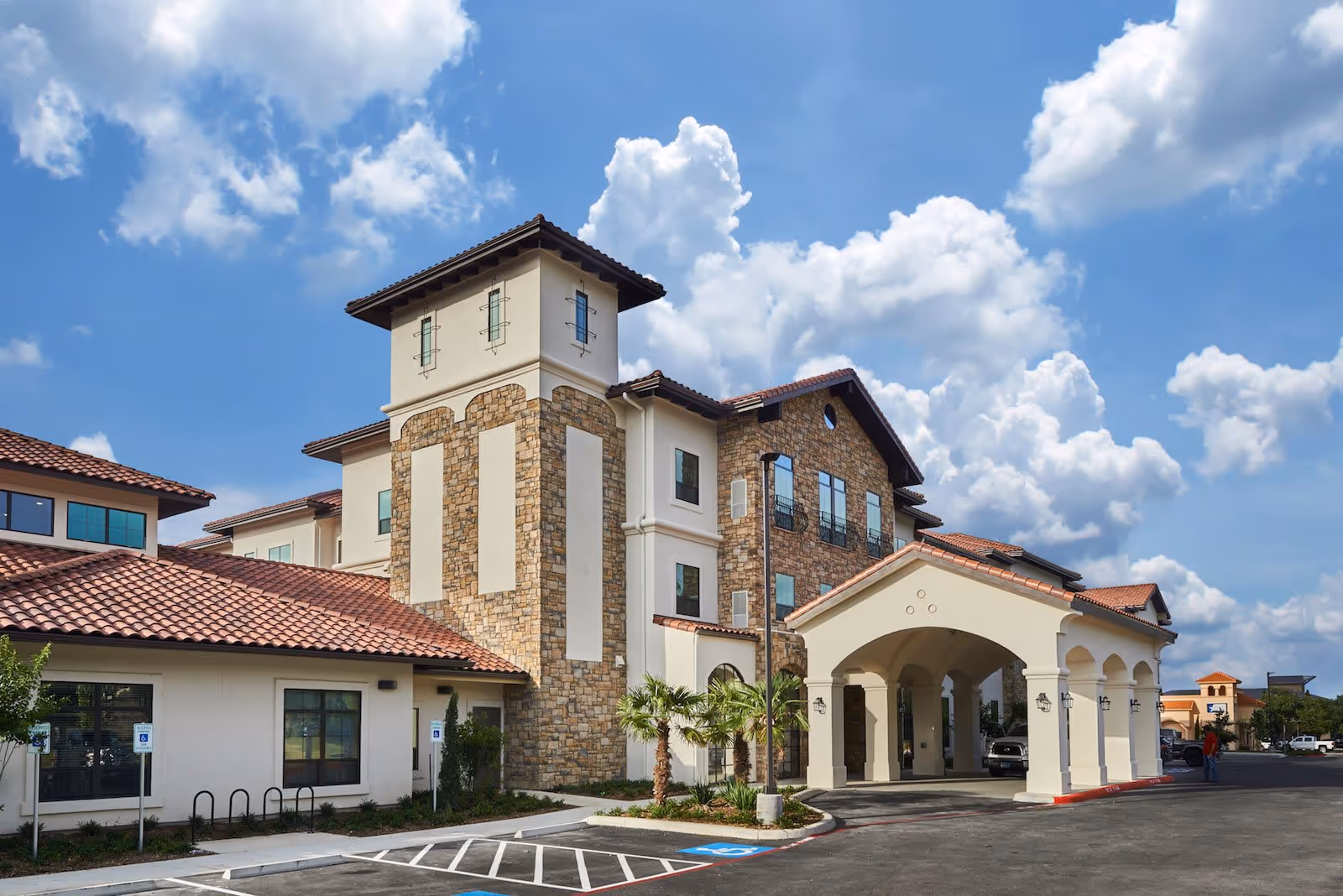 Exterior view of a multi-story assisted living facility with stone and stucco walls, red tile roofing, and a covered entrance. The building is surrounded by a parking lot with handicap parking spaces and small palm trees. The sky is blue with scattered clouds.