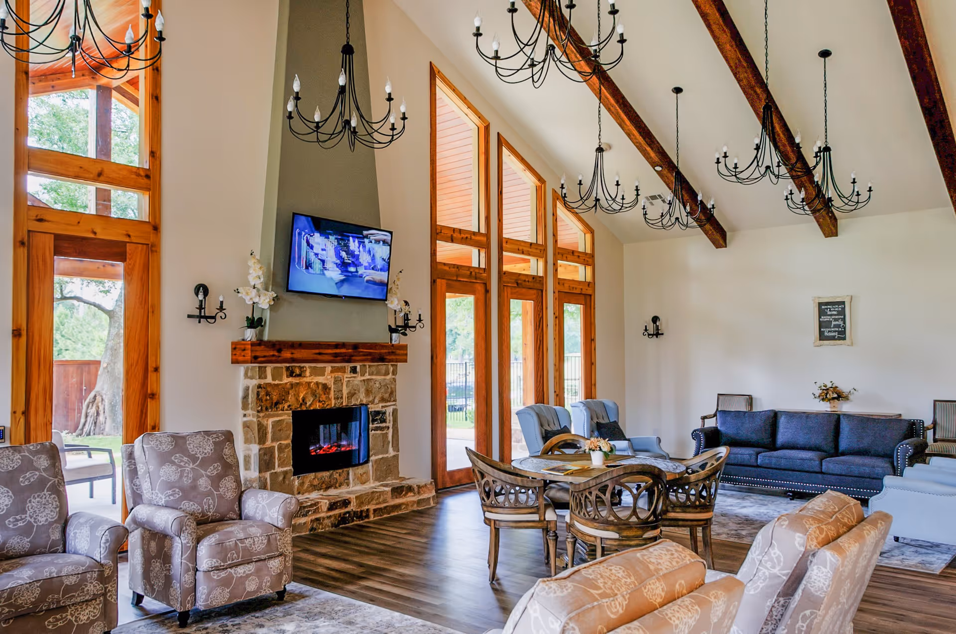 A spacious living room with high vaulted ceilings featuring exposed wooden beams and multiple black chandeliers. The room has large wooden-framed windows and doors letting in natural light. There is a stone fireplace with a mounted flat-screen TV above it, flanked by two white orchid plants. The seating includes patterned armchairs, a dark blue sofa, light blue armchairs, and a round wooden table with chairs. The floor is a combination of wood and area rugs, and the walls are painted white.