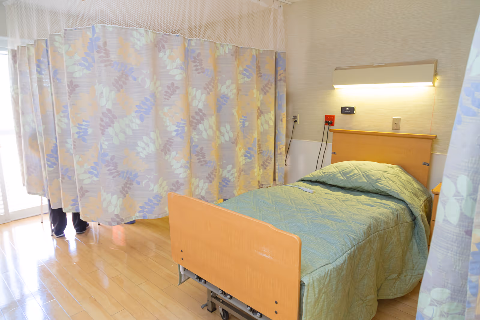 A hospital-style bed with a green quilted blanket in a room with light-colored walls and wooden flooring. A floral-patterned privacy curtain is partially drawn around the bed. There is a wall-mounted light above the bed and some electrical outlets and switches on the wall behind the headboard.