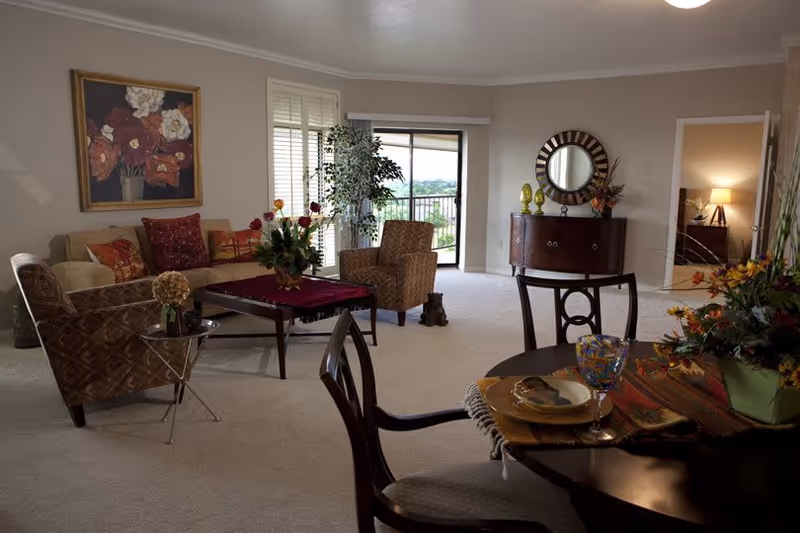 A cozy living room with beige carpet and walls, featuring a beige sofa with red and patterned cushions, two patterned armchairs, a coffee table with a red cloth and flower arrangement, a round dining table set with plates and glasses, a sideboard with decorative items and a round mirror above it, and a doorway leading to a bedroom with a lit lamp. There is a large window with shutters and a sliding glass door opening to a balcony with a view of greenery outside.