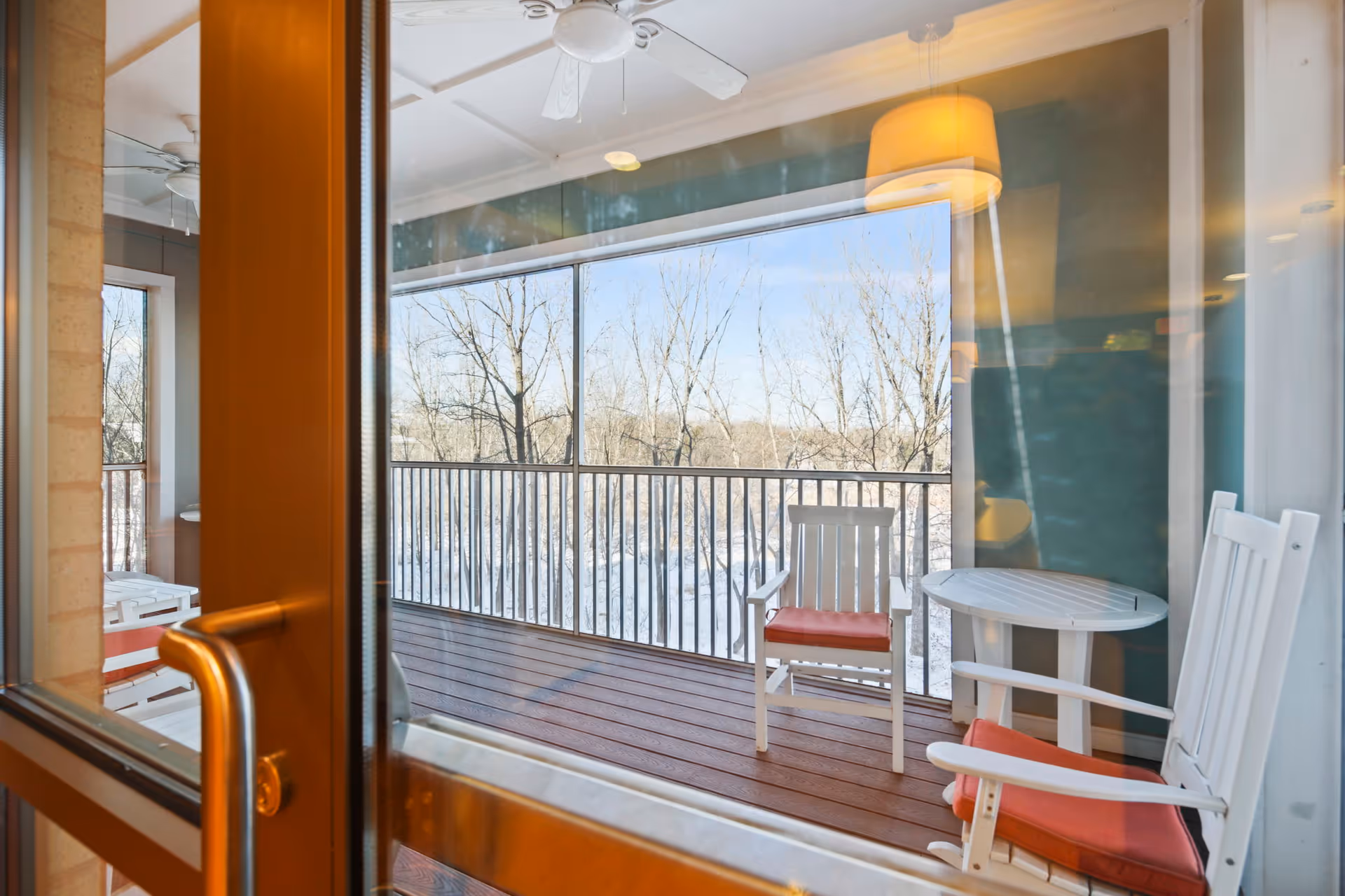 View through a glass door of a balcony with two white wooden chairs with red cushions and a small round white table. The balcony has a wooden floor and a black metal railing. Outside, leafless trees are visible under a clear blue sky.