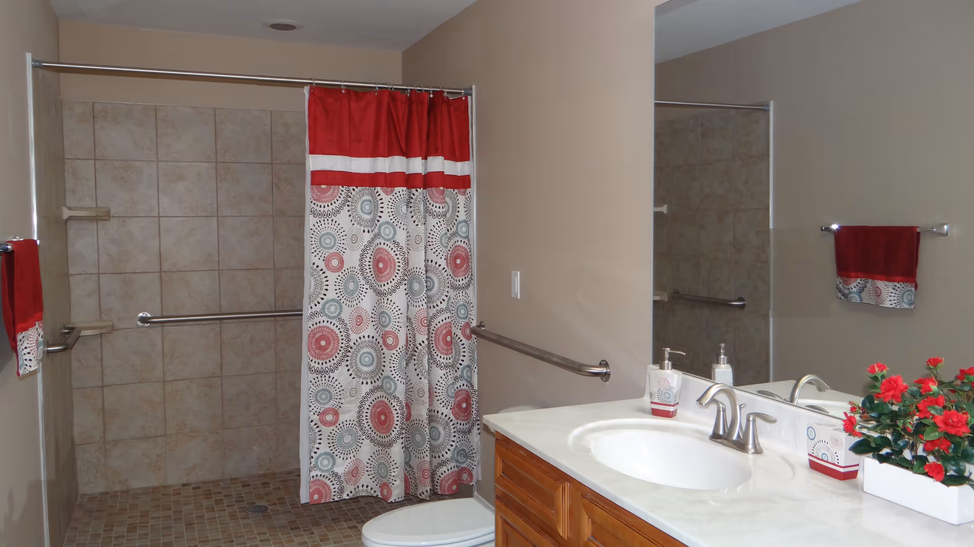 A bathroom with a tiled walk-in shower featuring a red, white, and patterned shower curtain. There are grab bars installed inside the shower and on the wall next to the sink. The sink countertop has a soap dispenser, a matching toothbrush holder, and a small potted plant with red flowers. A matching hand towel hangs on a towel rack beside the shower.