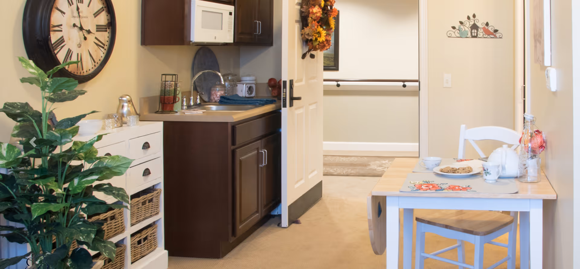 A cozy interior space featuring a small kitchen area with dark wood cabinets, a microwave, and a countertop with decorative items. To the right, there is a small wooden dining table set with a plate of cookies, cups, and a teapot. A white chair is tucked under the table. On the left side, there is a white storage unit with wicker baskets and a large green plant. A large wall clock is mounted above the storage unit. The room has beige walls and carpeted flooring, with a door leading to another room in the background.