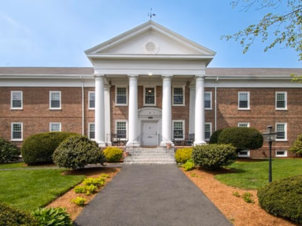 Front exterior of a two-story brick building with a white-columned portico, central entrance, and landscaped walkway.