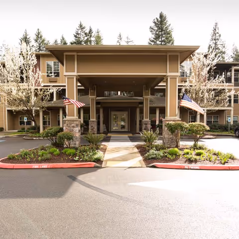 Entrance of a senior living facility with a covered drop-off area, two American flags on either side, landscaped greenery, and a three-story building in the background surrounded by trees.
