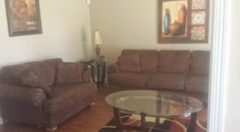 A living room with two brown upholstered sofas, a glass coffee table with a dark wooden frame, a patterned area rug, a small side table with a lamp, and framed artwork on the walls.