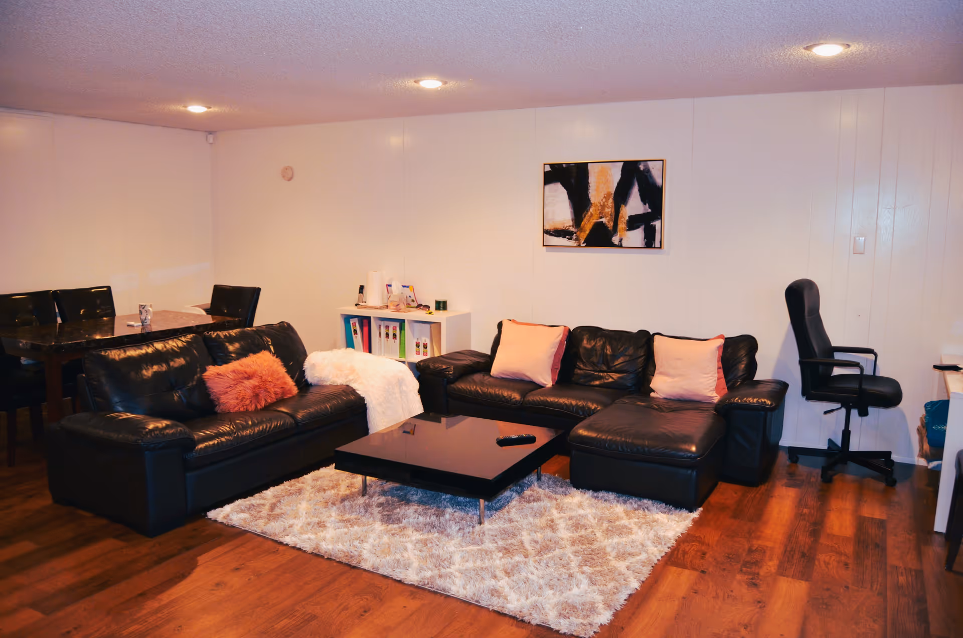 A cozy living room with two black leather sofas adorned with pink and white cushions, a fluffy white rug, and a black coffee table with a remote control on it. There is a small white shelf with books and items, a black office chair, and a dining table with chairs in the background. The walls are white with a modern abstract painting hanging above one sofa.