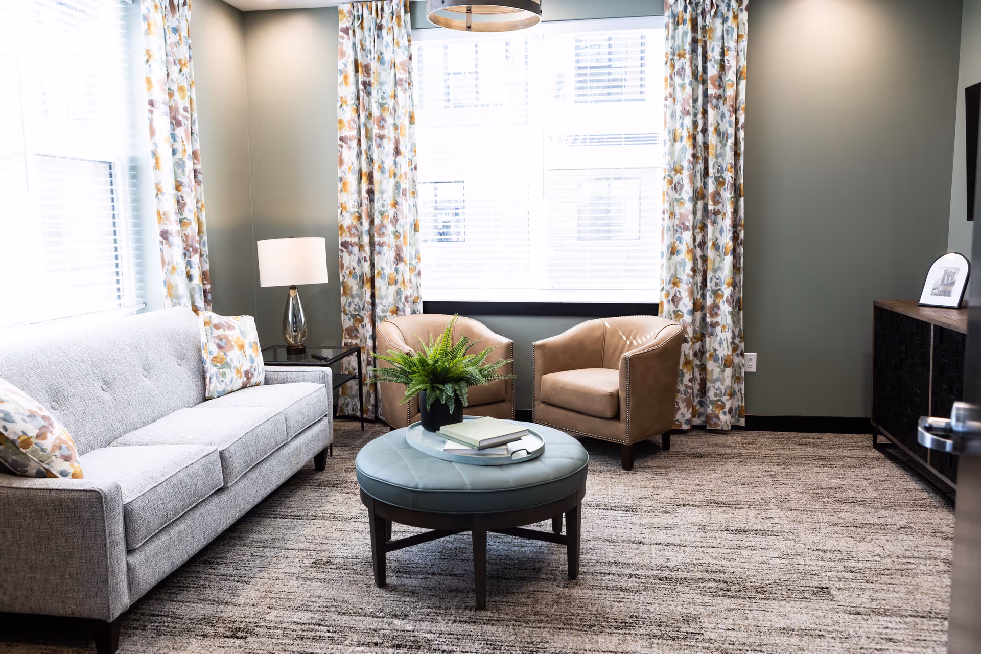A bright living room with a gray sofa adorned with floral pillows, two tan armchairs, a round blue ottoman with books and a plant on top, floral curtains framing large windows, a side table with a lamp, and a dark wooden cabinet with a framed photo.