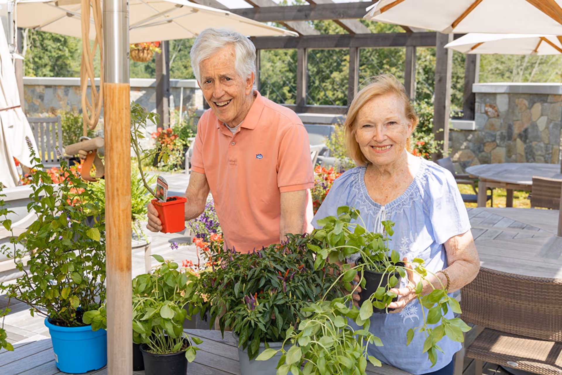 An elderly man and woman smiling and holding potted plants while gardening on a patio with tables, chairs, and umbrellas in the background.