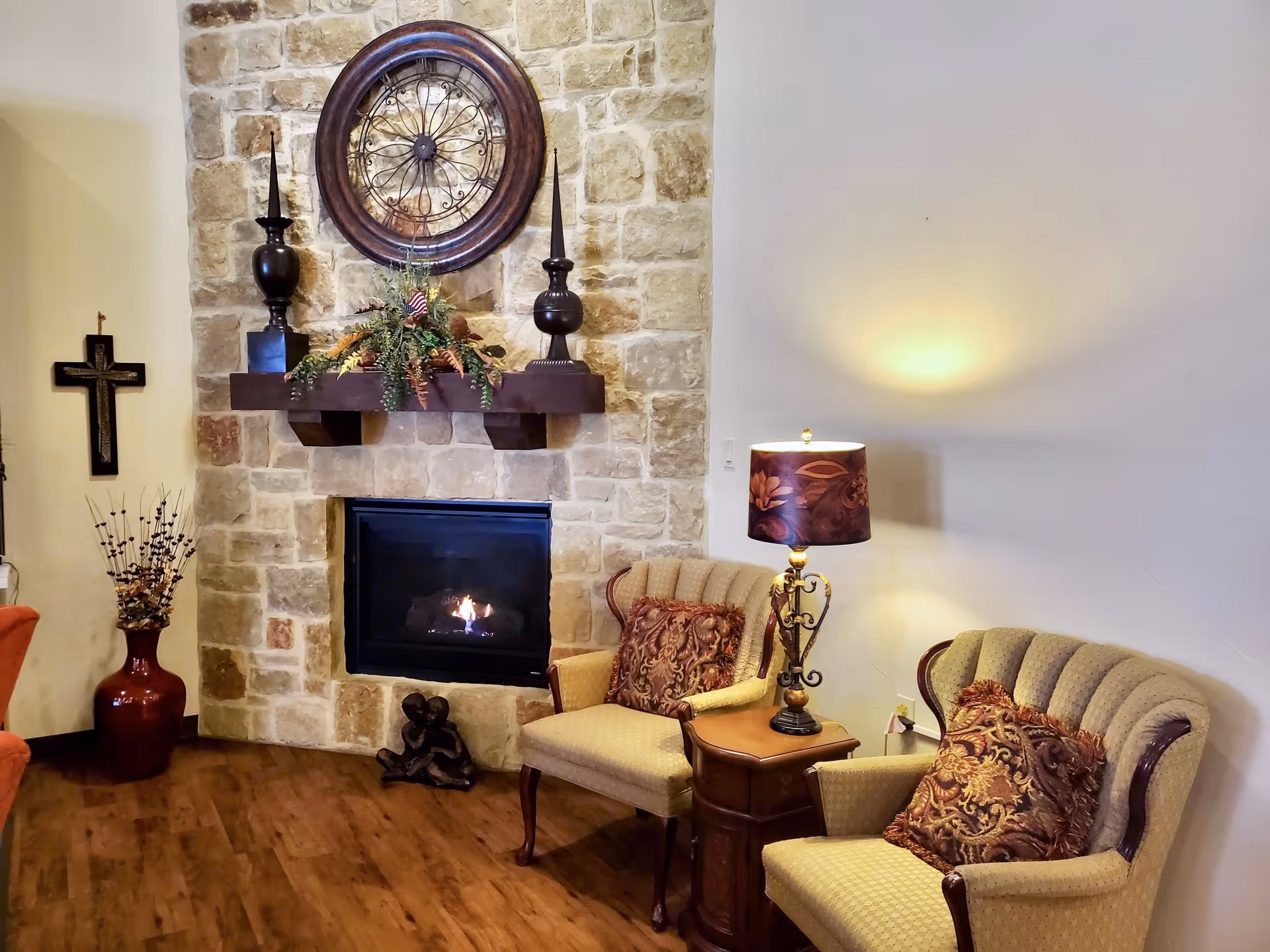 A cozy living room area with a stone fireplace, a wooden mantel decorated with two black candlesticks and a floral arrangement. Above the mantel is a large round decorative wall clock. Two upholstered armchairs with patterned cushions are placed next to a wooden side table with an ornate lamp. A large red vase with decorative branches and a wall cross are visible on the left side.