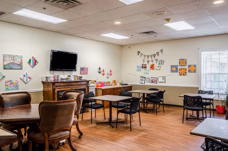 A bright communal activity/dining room with multiple tables and chairs, a wall-mounted TV, and colorful artwork on the walls.