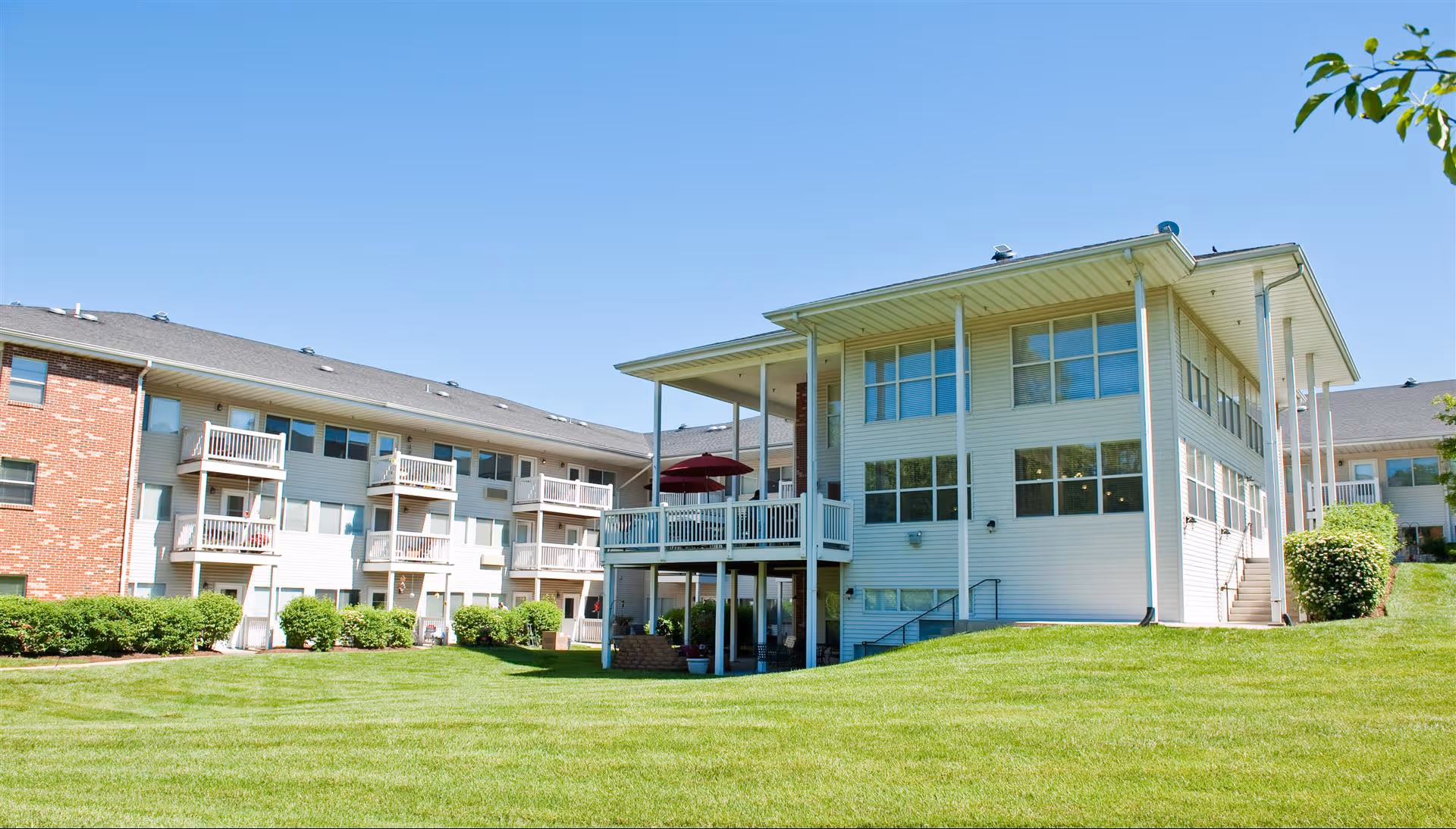 Exterior view of a senior living facility building with multiple balconies, large windows, and a covered patio area. The building is surrounded by a well-maintained green lawn and bushes under a clear blue sky.