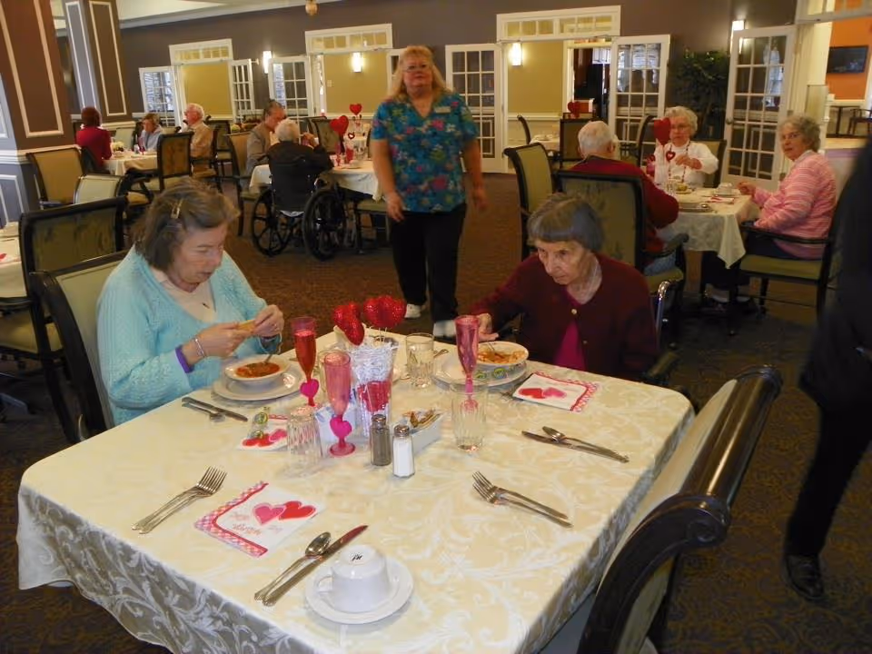 Several elderly individuals seated at tables in a dining room, eating and socializing. The tables are covered with white tablecloths and decorated with red heart-themed decorations and napkins. A staff member in a blue floral shirt is walking between the tables. The room has a warm, inviting atmosphere with multiple tables and chairs arranged for dining.