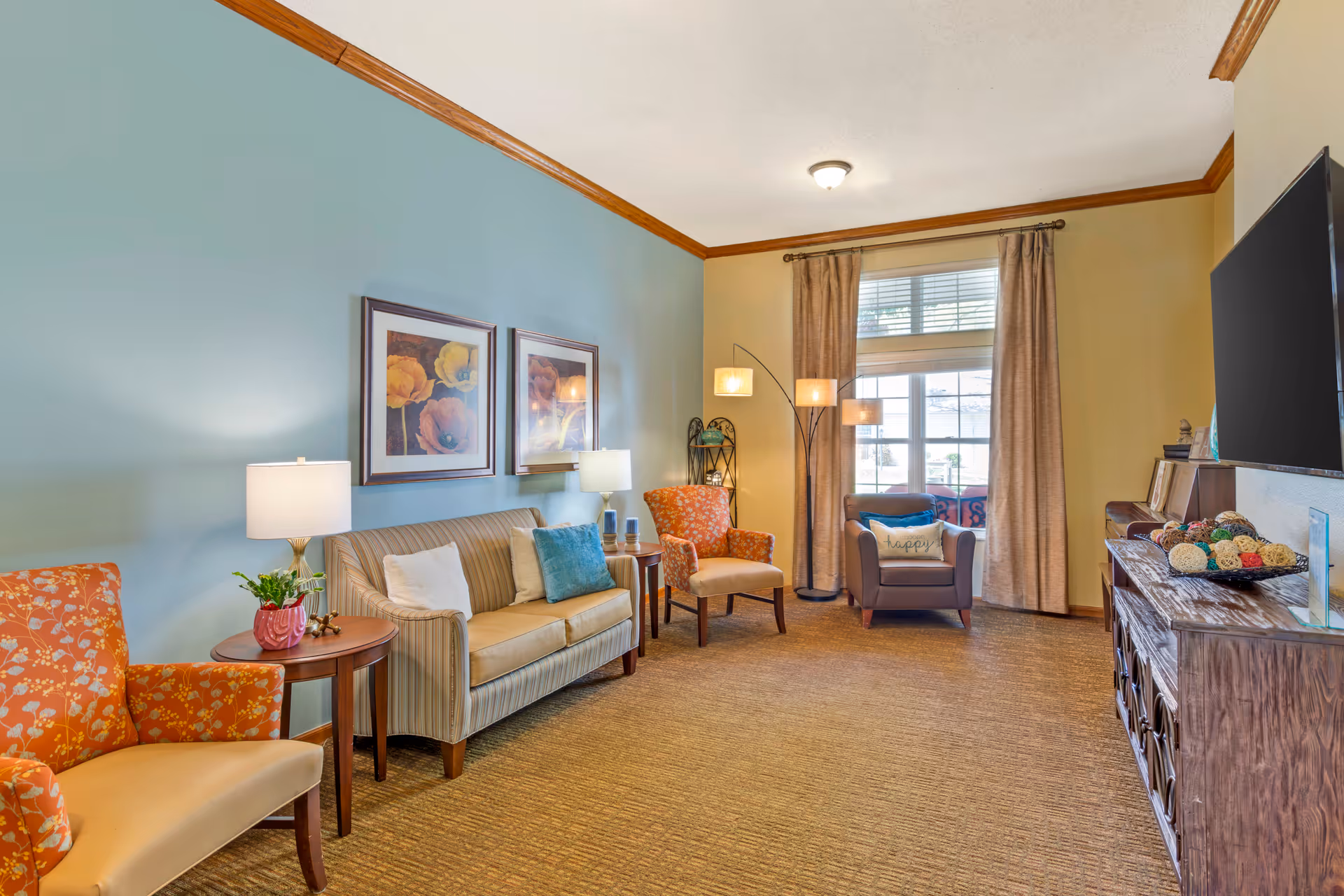 A cozy living room with a striped sofa, two orange floral armchairs, a brown armchair with a 'choose happy' pillow, a wooden side table with a lamp and plant, a floor lamp with three lights, two framed floral paintings on a blue accent wall, a large window with beige curtains, and a wooden TV stand with decorative items and a flat-screen TV mounted on the wall.