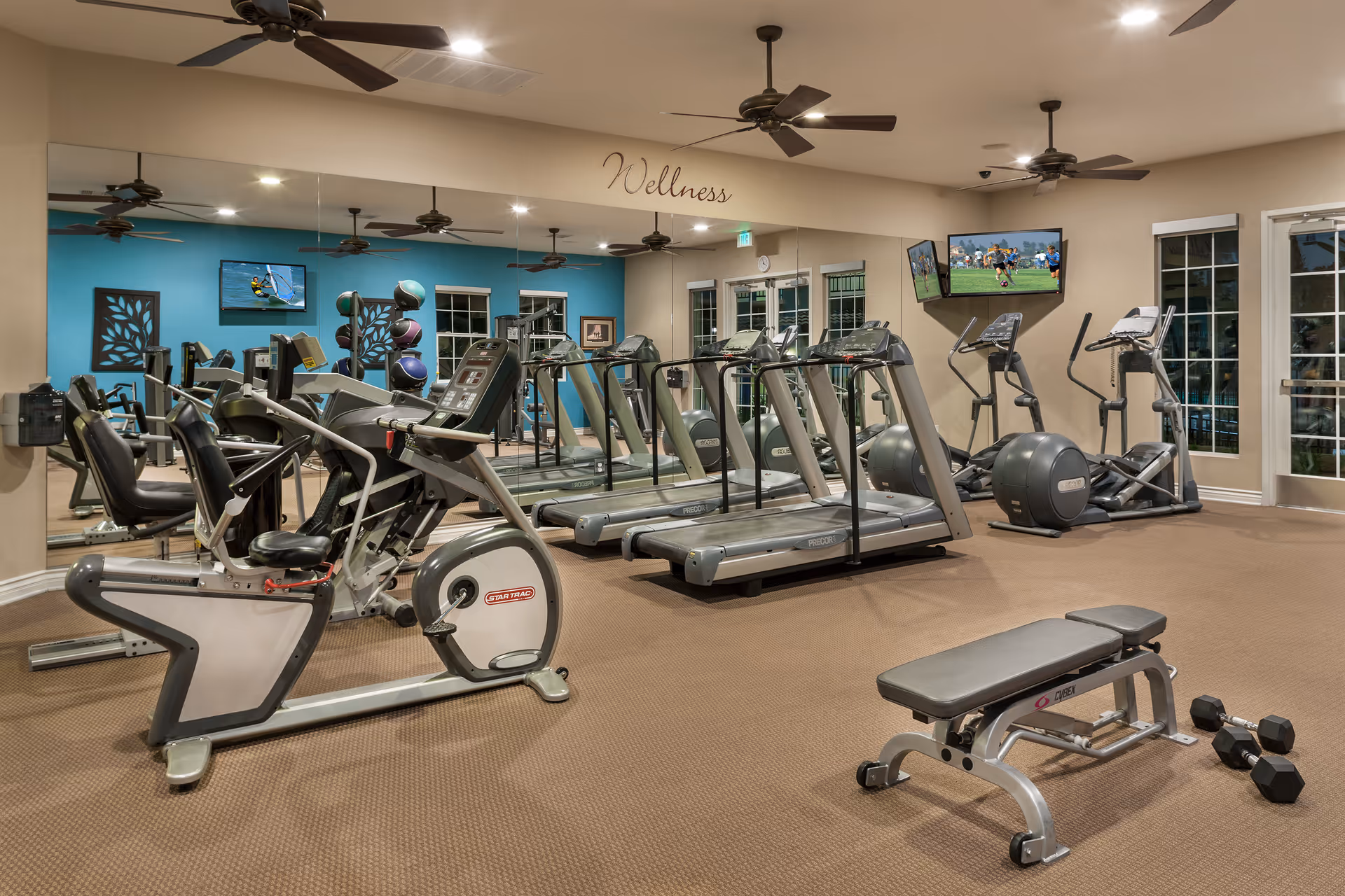Well-lit wellness/fitness room with treadmills, exercise bikes, a weight bench, dumbbells, mirrored wall and ceiling fans.