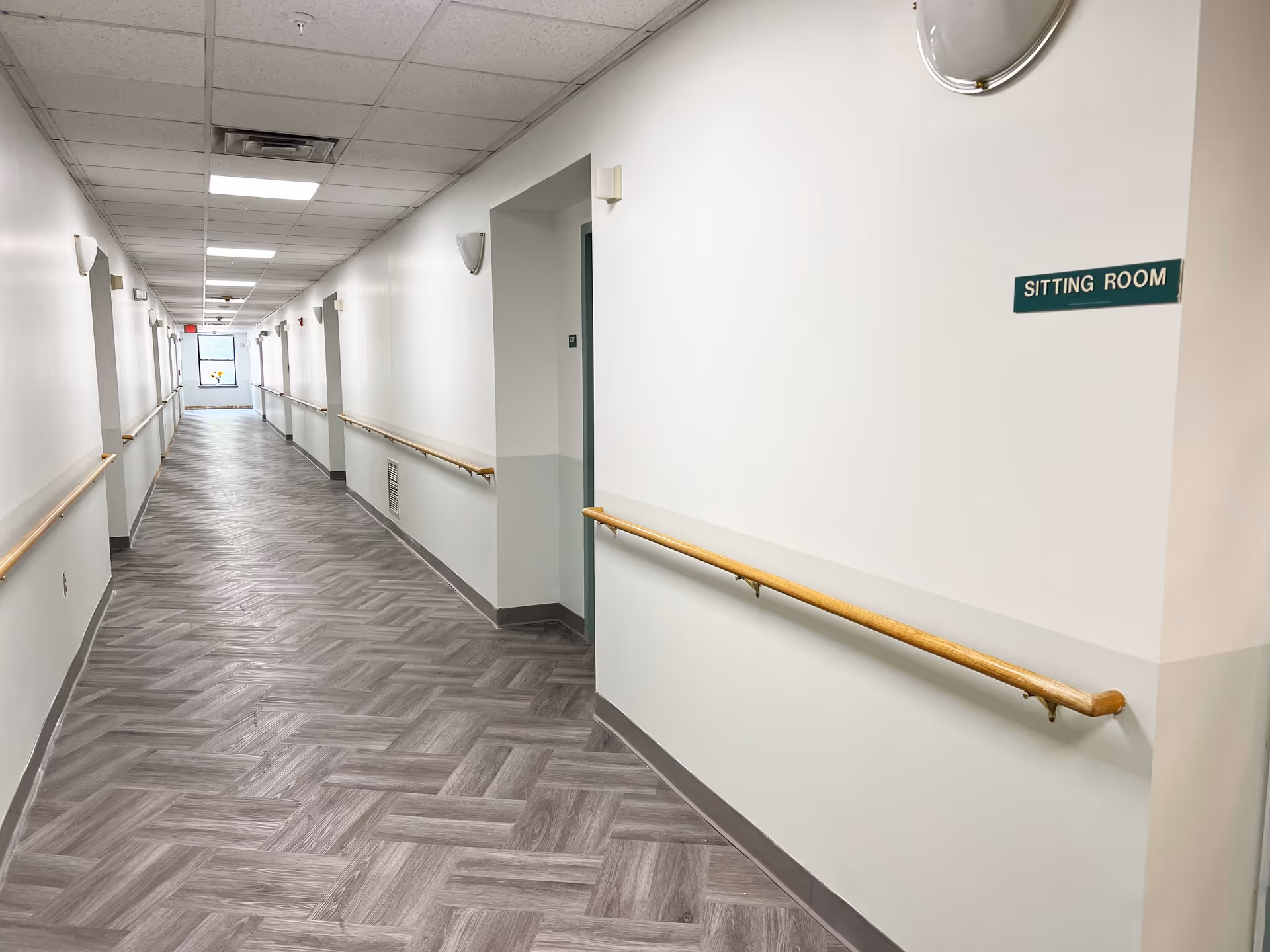 A long, empty hallway in a senior living facility with light-colored walls and wood-patterned flooring. Handrails run along both sides of the hallway, and there are several doors and wall-mounted lights. A small green sign on the right wall reads 'SITTING ROOM'.