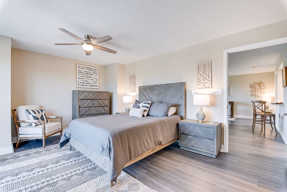 Bright furnished bedroom with a gray wood bed, matching nightstands and dresser, an armchair, ceiling fan, and a doorway leading to a dining area.