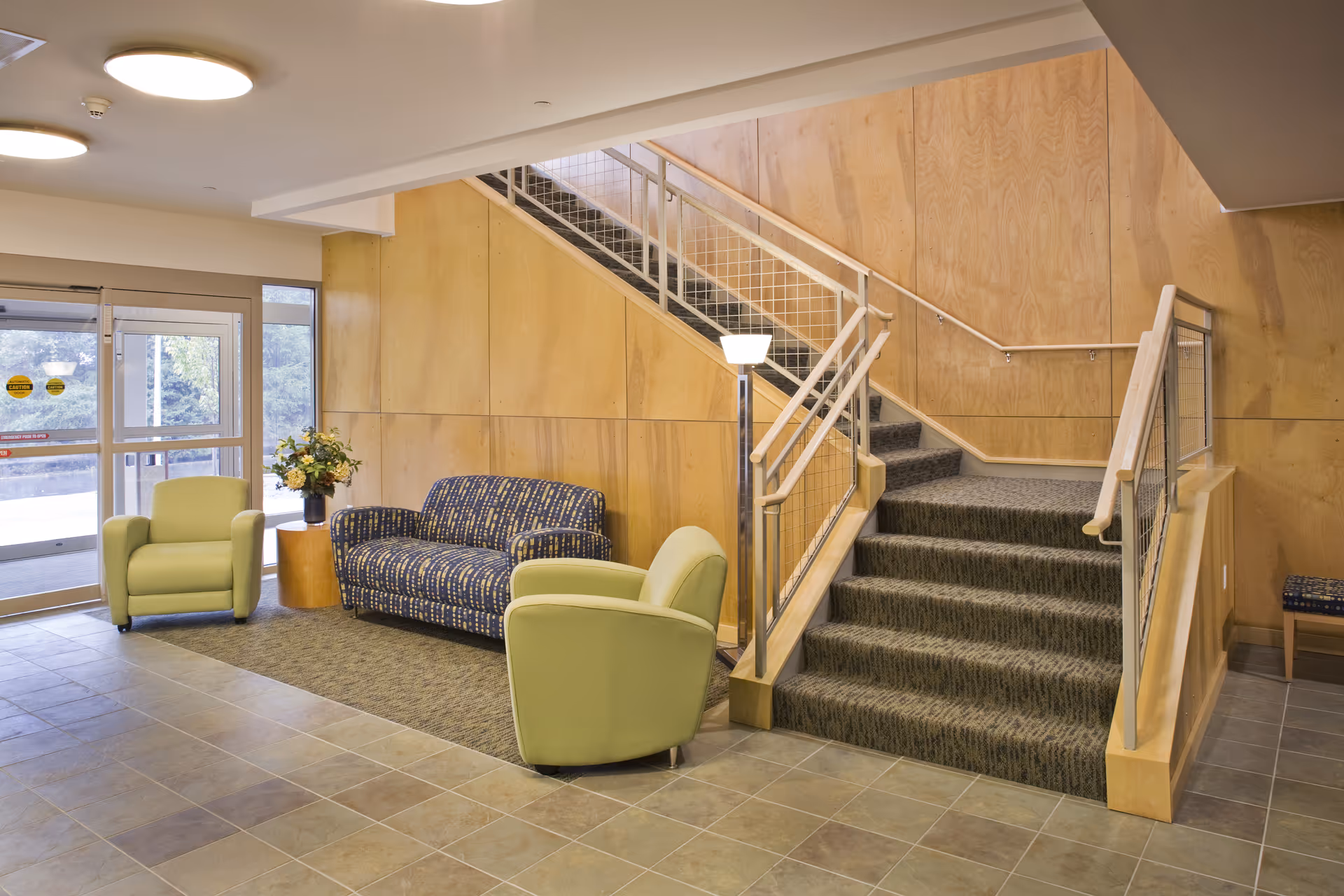 Interior view of a retirement residence lobby area with a staircase. The space features a patterned blue sofa, two light green armchairs, a small wooden side table with a flower arrangement, and large windows near the entrance door. The walls are wooden paneled and the floor is tiled.