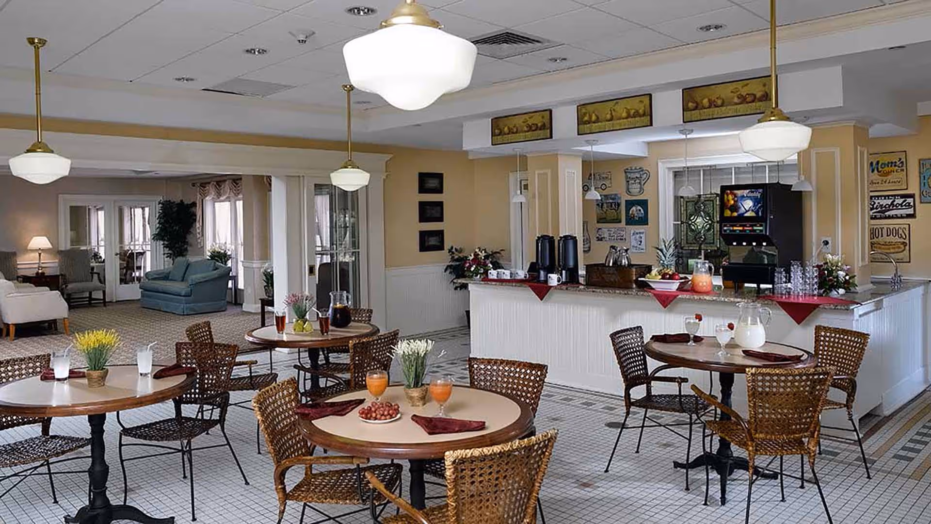 A cozy dining area in a senior living facility with round tables and wicker chairs. Each table has drinks and small decorative plants. In the background, there is a counter with coffee dispensers, pitchers, and a soda machine. The room has warm lighting with hanging pendant lights and a comfortable seating area with armchairs and a sofa visible through an open doorway.