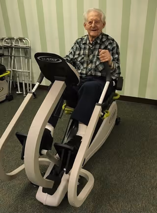An elderly man wearing a plaid shirt and dark pants is seated on a NuStep exercise machine in a room with striped green wallpaper and carpeted floor. There are walkers visible in the background.