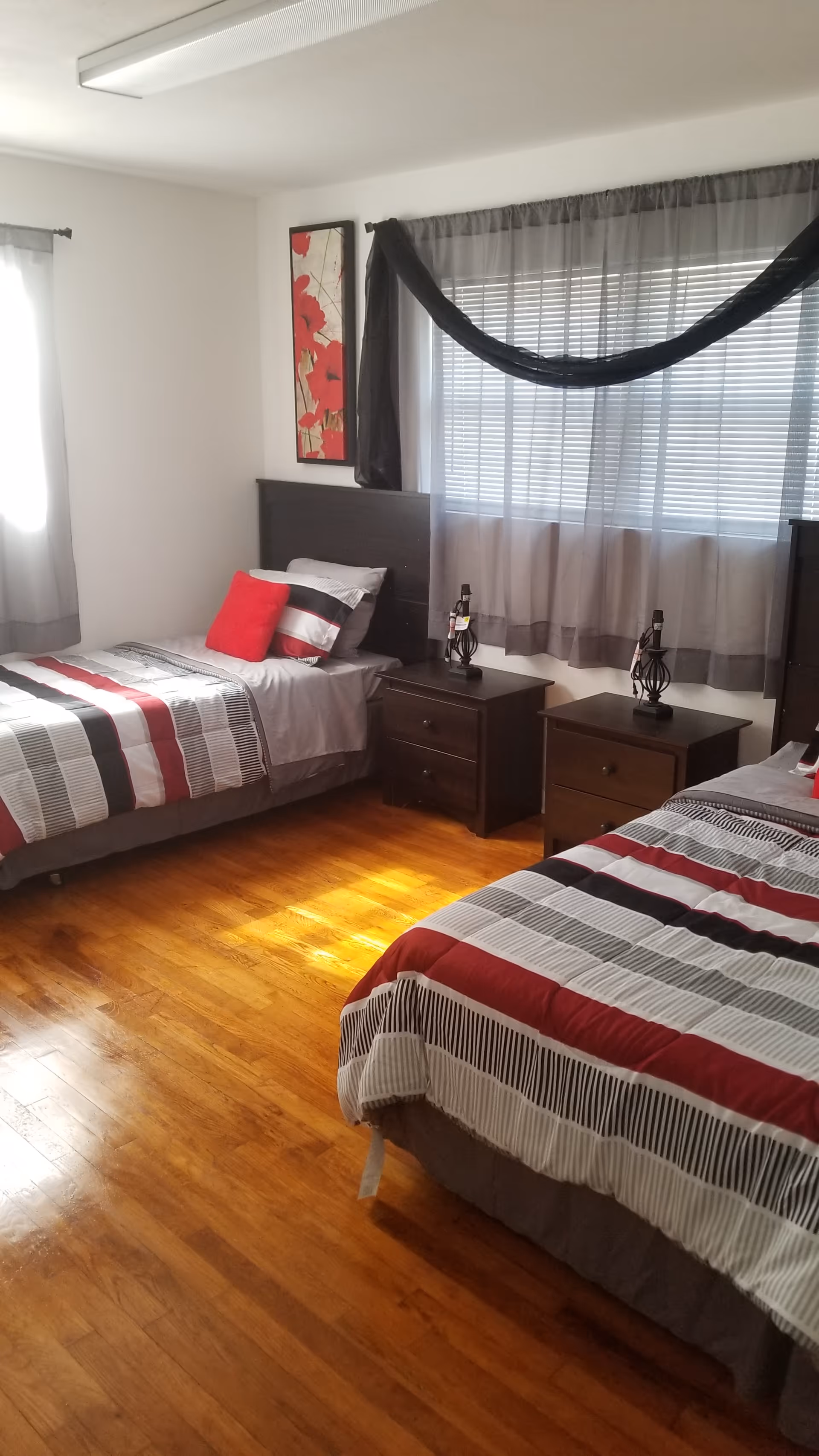 A bedroom with two single beds featuring matching striped bedding in red, black, white, and gray. Each bed has a pillow and a red accent pillow. Between the beds are two dark wood nightstands with decorative lamps. A window with gray curtains and blinds is above the nightstands. The room has hardwood floors and a piece of wall art with red flowers.