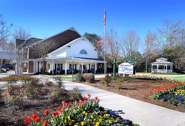 Exterior view of Belmont Village Senior Living Buckhead facility with a white building featuring a covered porch, an American flag on a flagpole, a garden with colorful flowers including red tulips, and a white gazebo in the background under a clear blue sky.
