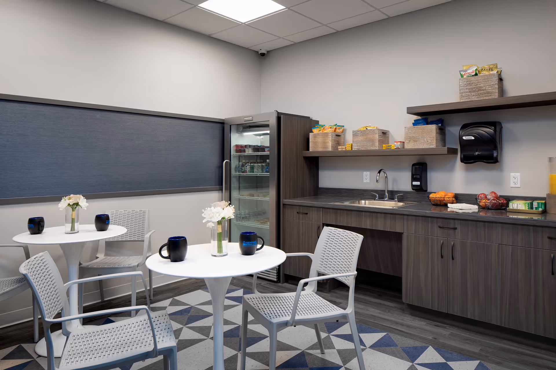Small kitchenette and seating area with round tables and chairs, a sink and countertop with snacks and a glass-front refrigerator.