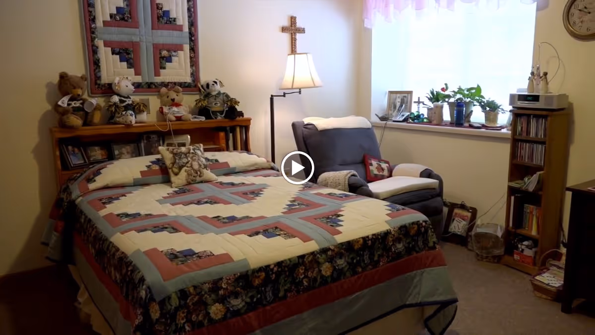 A cozy bedroom with a bed covered in a patterned quilt, several stuffed animals on the headboard, a cushioned armchair with a throw blanket and pillow, a floor lamp, a window with sheer curtains, and a small wooden shelf holding plants, framed photos, and CDs.