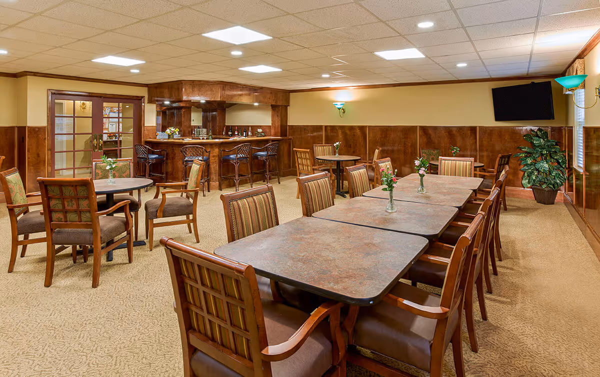 Community dining room with long tables and chairs, small round tables, a bar area and a wall-mounted TV.