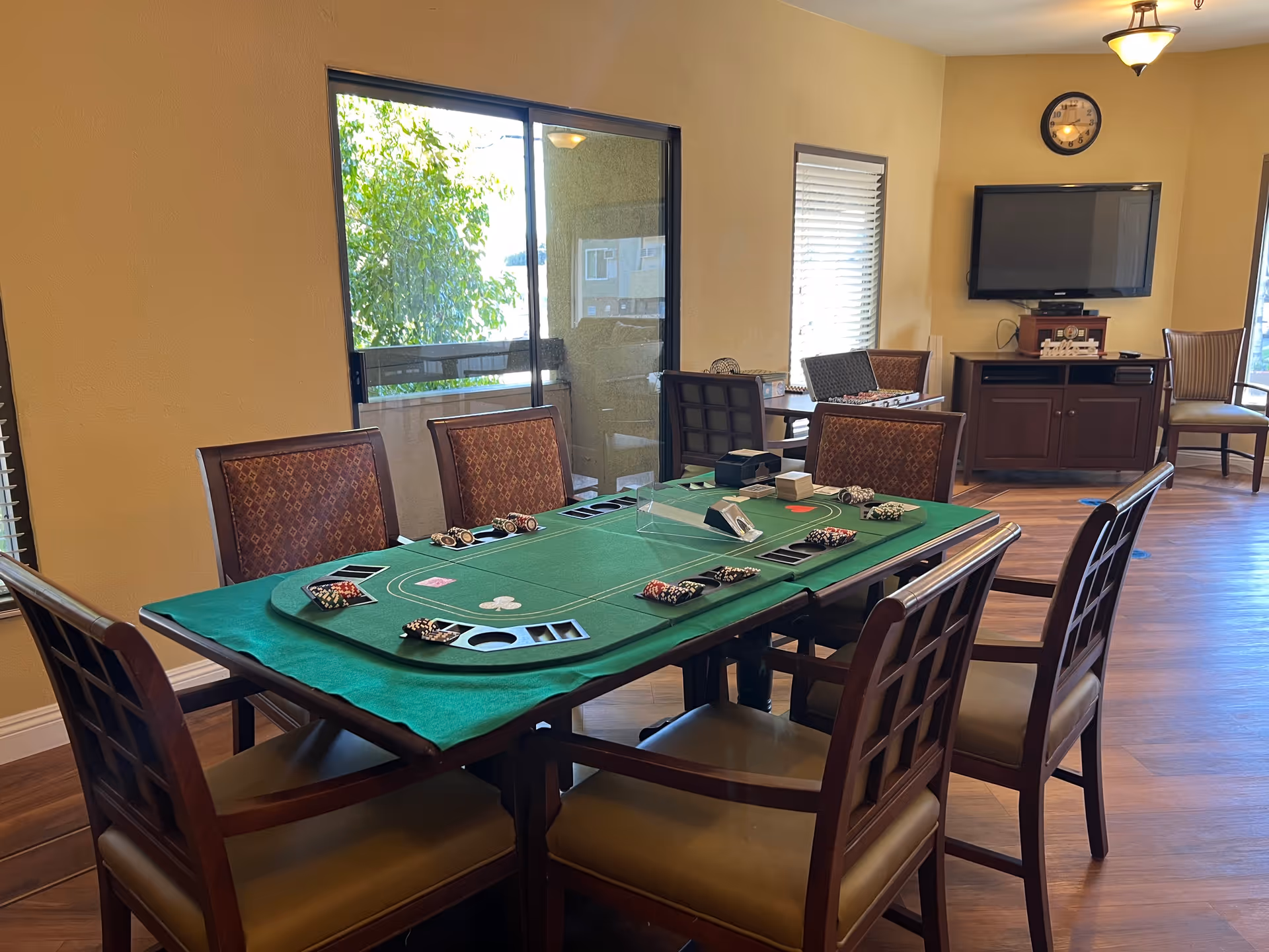 A room with a poker table covered in a green felt cloth, poker chips, and cards set up for a game. The room has wooden chairs around the table, a wall-mounted TV, a clock above the TV, and windows letting in natural light.