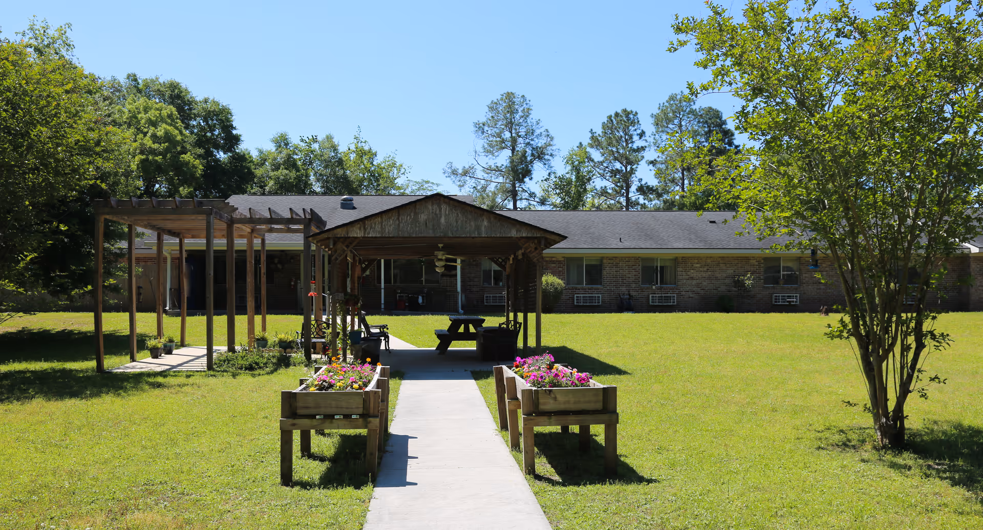 A grassy courtyard with a central paved path leading to a wooden gazebo and picnic table, flanked by raised flowerbeds and a single-story brick building in the background.