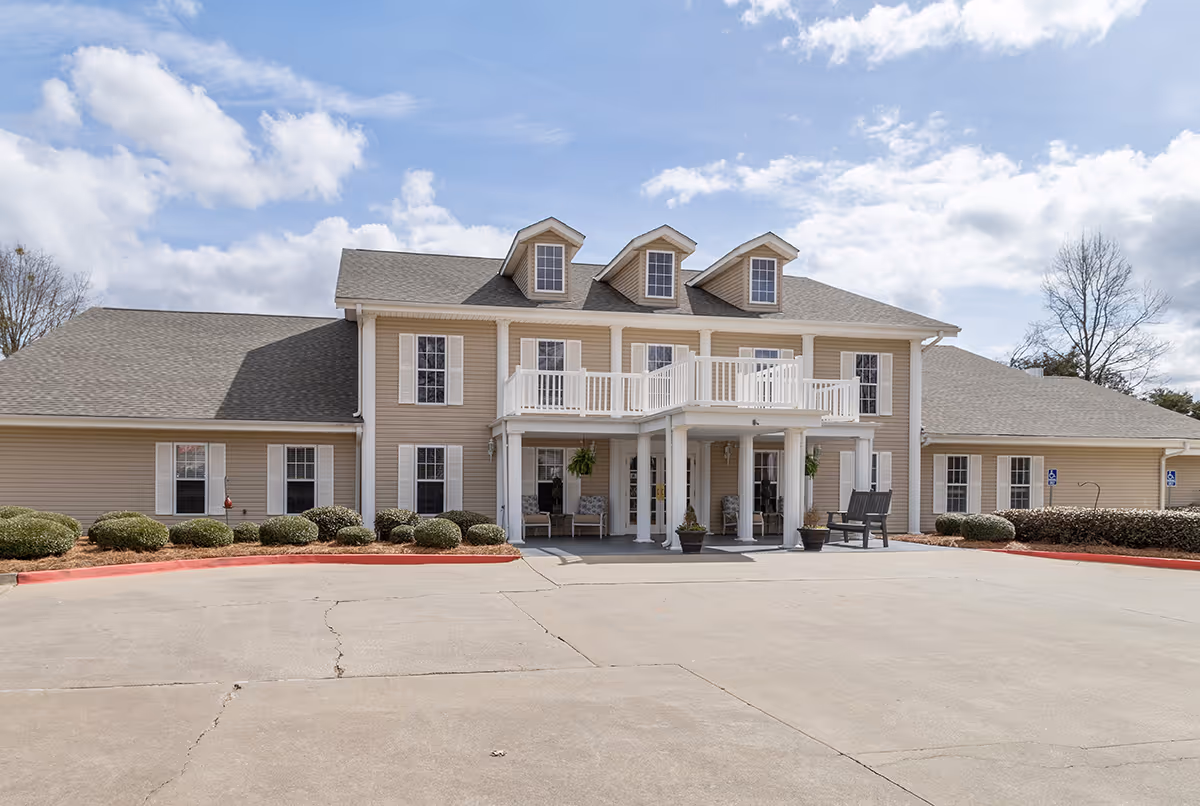 Front exterior view of a two-story beige building with white trim and a covered entrance supported by columns. The building has three dormer windows on the roof and a balcony above the entrance. Shrubs line the front of the building and a concrete driveway is in the foreground under a partly cloudy sky.