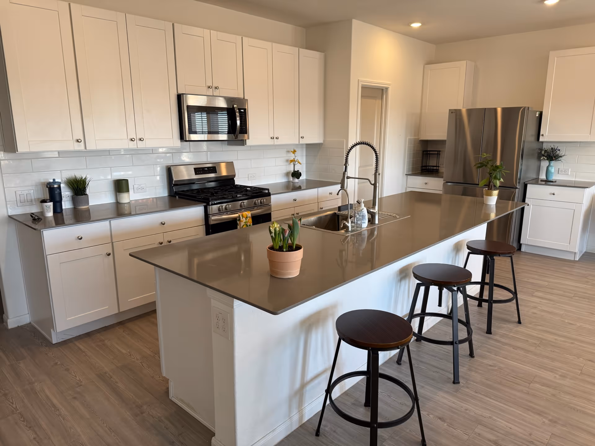 A modern kitchen with white cabinets, stainless steel appliances including a microwave, stove, and refrigerator. There is a large island with a gray countertop, a sink with a tall faucet, and three dark wooden stools. Several small potted plants are placed on the countertops and island. The floor is light wood, and the walls are white.