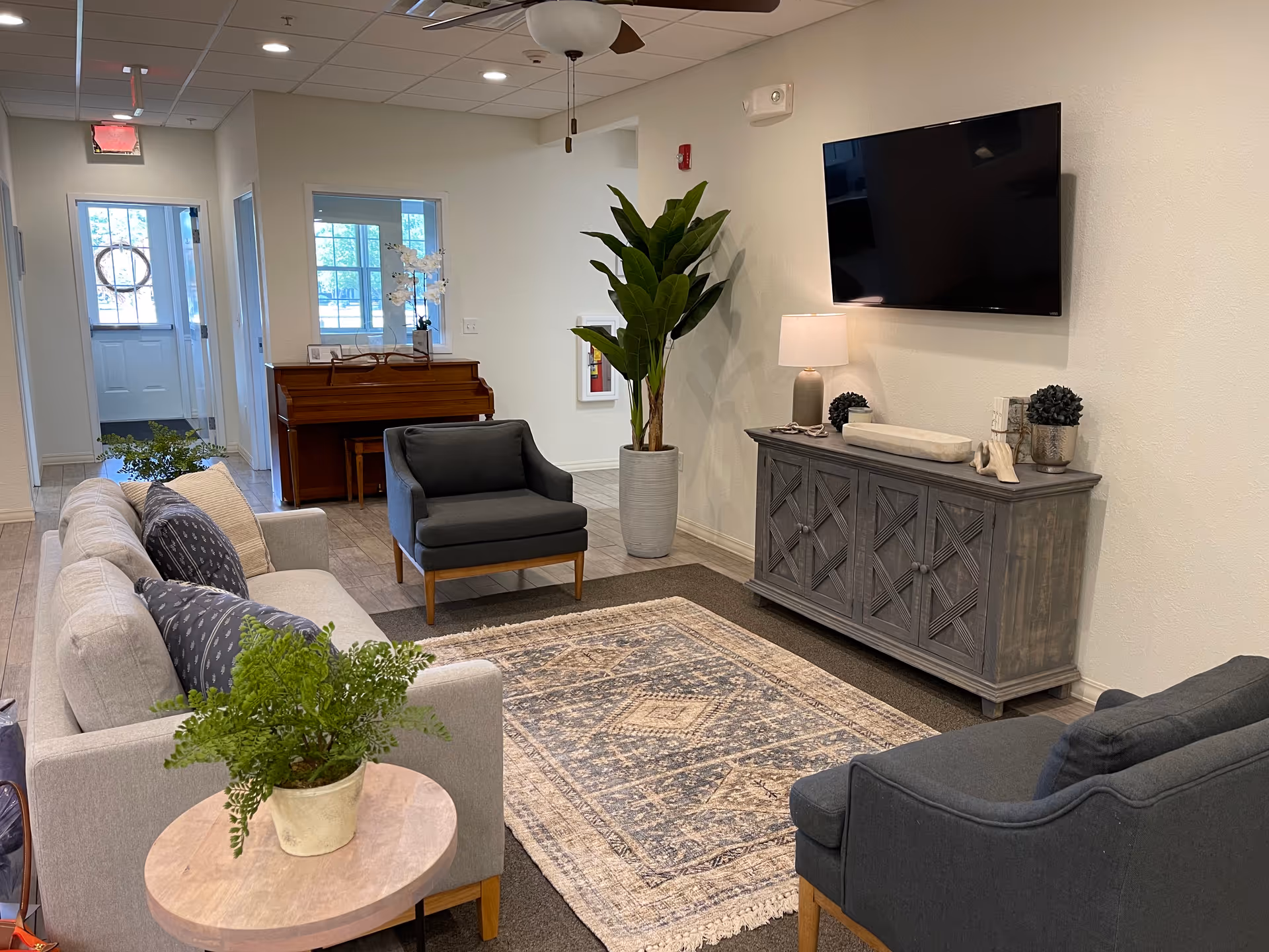 A cozy living room area in a memory care facility featuring a light gray sofa with patterned pillows, two dark gray armchairs, a wooden side table with a potted plant, a large patterned area rug, a wooden cabinet with decorative items and a table lamp, a flat-screen TV mounted on the wall, a tall potted plant, and a piano in the background near a hallway with natural light coming through windows.